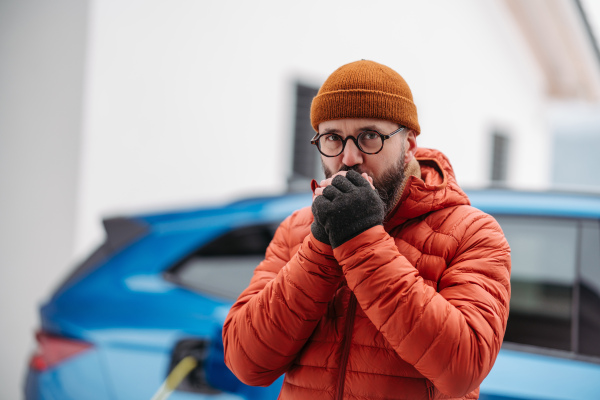 Man charging electric car during cold snowy day. Front view of hansome man feeling cold during freezing winter weather. Charging and driving electric vehicles during winter season.