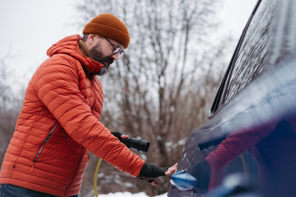 Man charging electric car during cold snowy day. Side view of hansome mature man putting charger in charging port during cold weather. Charging and driving electric vehicles during winter season.