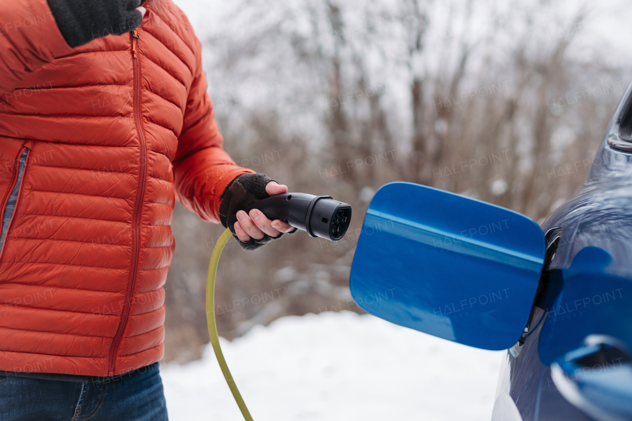 Close up of a charging electric car during cold snowy day, putting charger in charging port. Charging and driving electric vehicles during winter season.