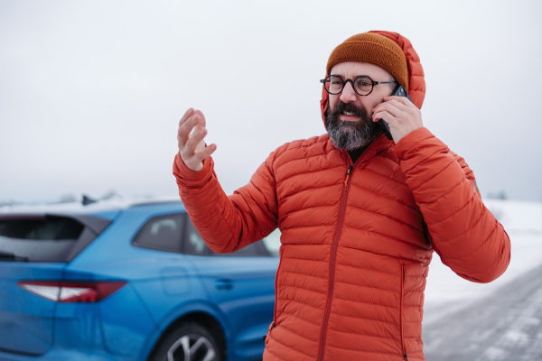 Angry man standing by electric car, battery run out of power before reaching destination. Man phone calling for help, waiting for the breakdown service car, tow truck.