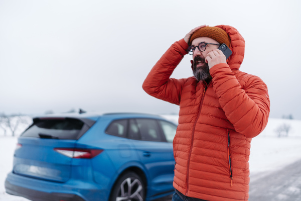 Angry man standing by electric car, battery run out of power before reaching destination. Man phone calling for help, waiting for the breakdown service car, tow truck.