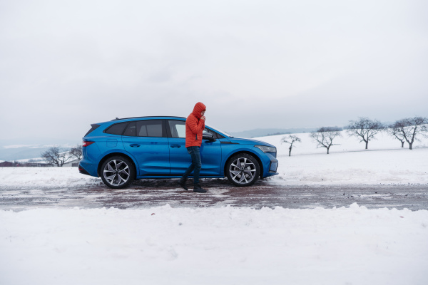 Angry man standing by electric car, battery run out of power before reaching destination, during snowy winter day. Frustrated man is cold, waiting for help, breakdown service car, tow truck.