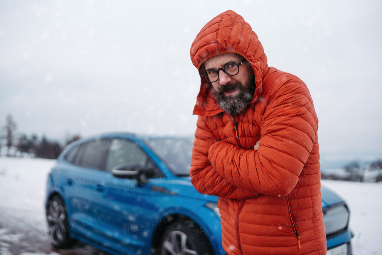 Angry man standing by electric car, battery run out of power before reaching destination, during snowy winter day. Frustrated man is cold, waiting for help, breakdown service car, tow truck.