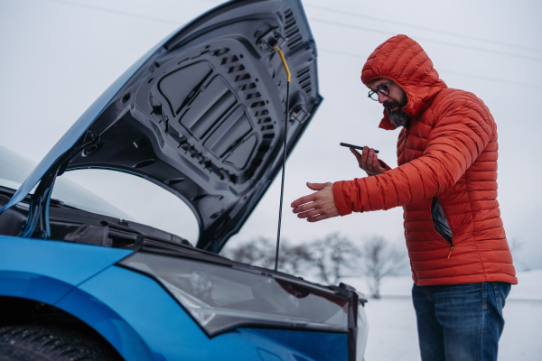 Angry man standing by electric car, battery run out of power before reaching destination. Man phone calling for help, ooking undre the car hood, waiting for the breakdown service car, tow truck.