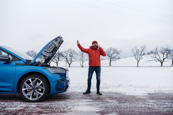 Angry man standing by electric car, battery run out of power before reaching destination. Man phone calling for help, ooking undre the car hood, waiting for the breakdown service car, tow truck.