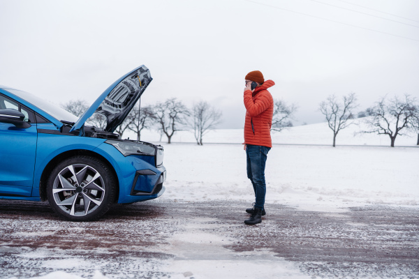 Angry man standing by electric car, battery run out of power before reaching destination. Man phone calling for help, ooking undre the car hood, waiting for the breakdown service car, tow truck.