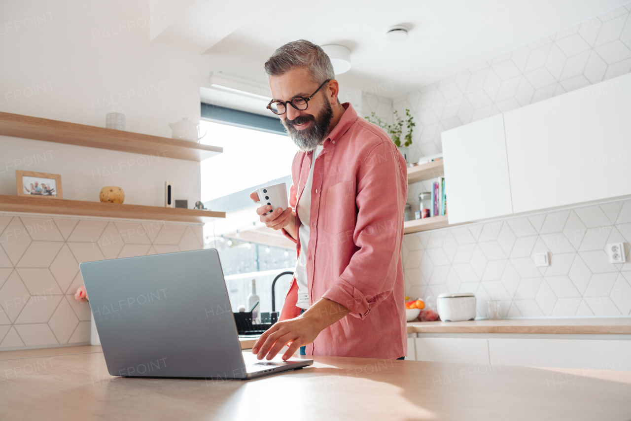 Mature man in pink shirt working from home on laptop, standing by kitchen island, with cup of coffee, holding smartphone. Concept of remote work from home. Hygge at work.