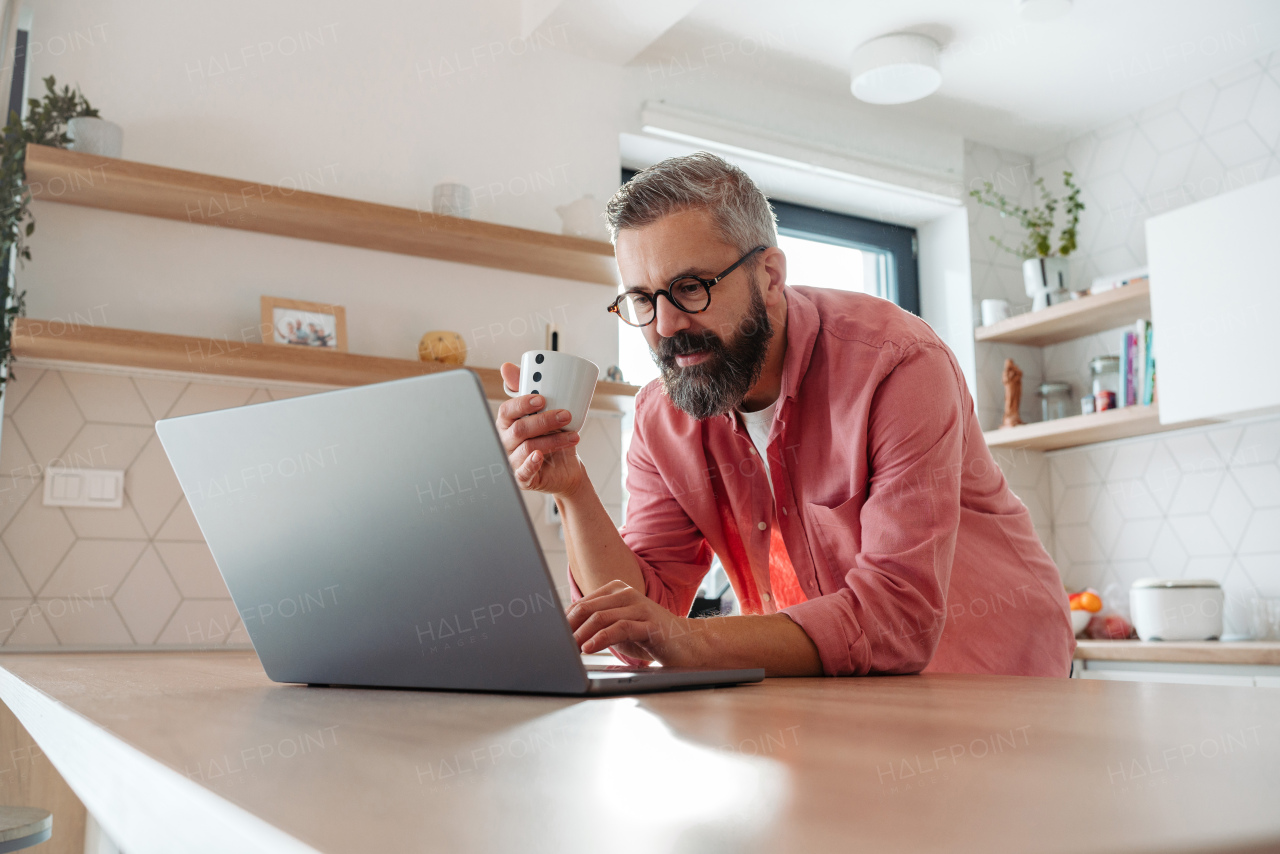 Mature man in pink shirt working from home on laptop, standing by kitchen island, with cup of coffee, holding smartphone. Concept of remote work from home. Hygge at work.