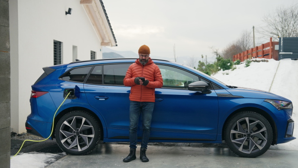 Mature man charging electric car during cold snowy day, using electric vehicle charging app, checking energy consumption, battery life on smart phone. Charging and driving electric vehicles during winter season.