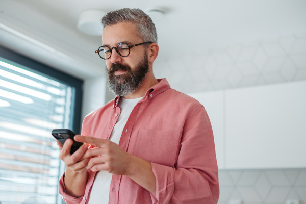 Portrait of man in pink shirt working from home holding smartphone, scrolling, standing by kitchen island. Concept of remote work from home. Hygge at work.