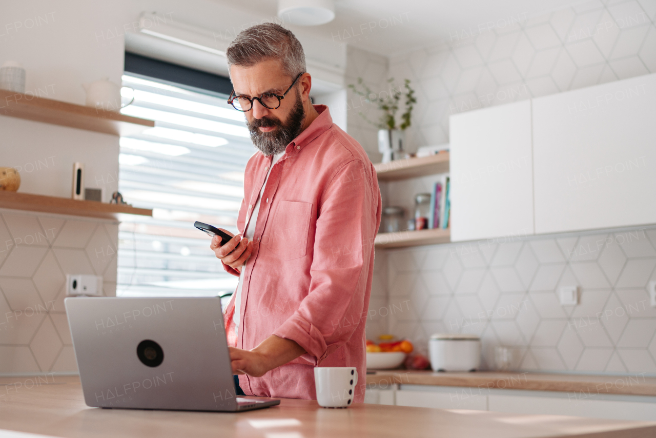 Mature man in pink shirt working from home on laptop, standing by kitchen island, with cup of coffee, holding smartphone. Concept of remote work from home. Hygge at work.