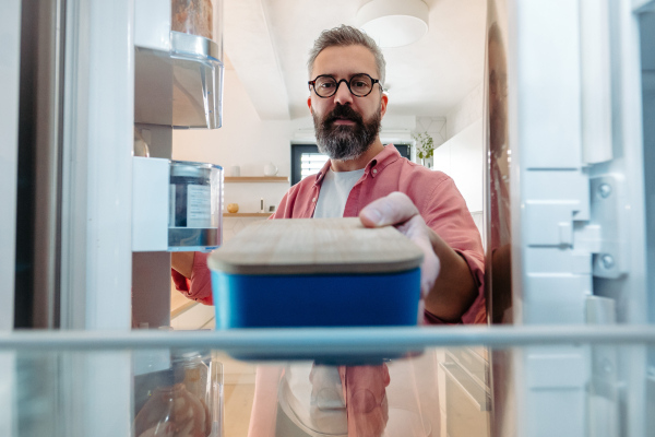 Handsome man putting lunchbox in fridge. Taking out lunch from fridge, eating leftovers. Do not trow food away, reducting food waste.