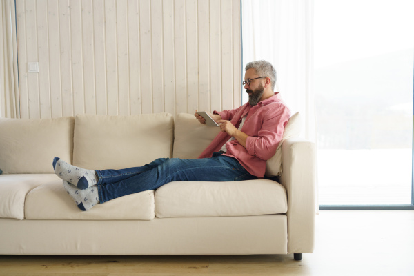 Man using smart thermostat, adjusting, lowering heating temperature at home. Man watching tv show on tablet, video streaming services with movies and tv shows online. Full length.