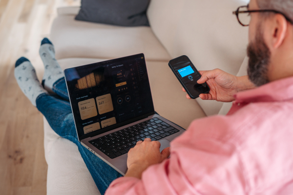 Man with notebook and smartphone sitting on sofa, online banking, internetbanking at home throught bank's website and app. Paying with smartphone online, contactless payments by mobile phone, virtual or digital wallets.