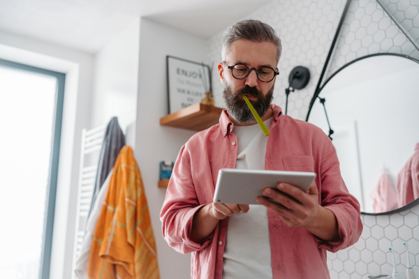 Man shopping online while brushing his teeth in bathroom. Holding tablet, scrolling and buying in eshop with toothbrush in mouth.