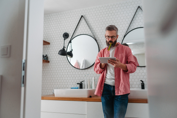 Man shopping online while brushing his teeth in bathroom. Holding tablet, scrolling and buying in eshop with toothbrush in mouth.