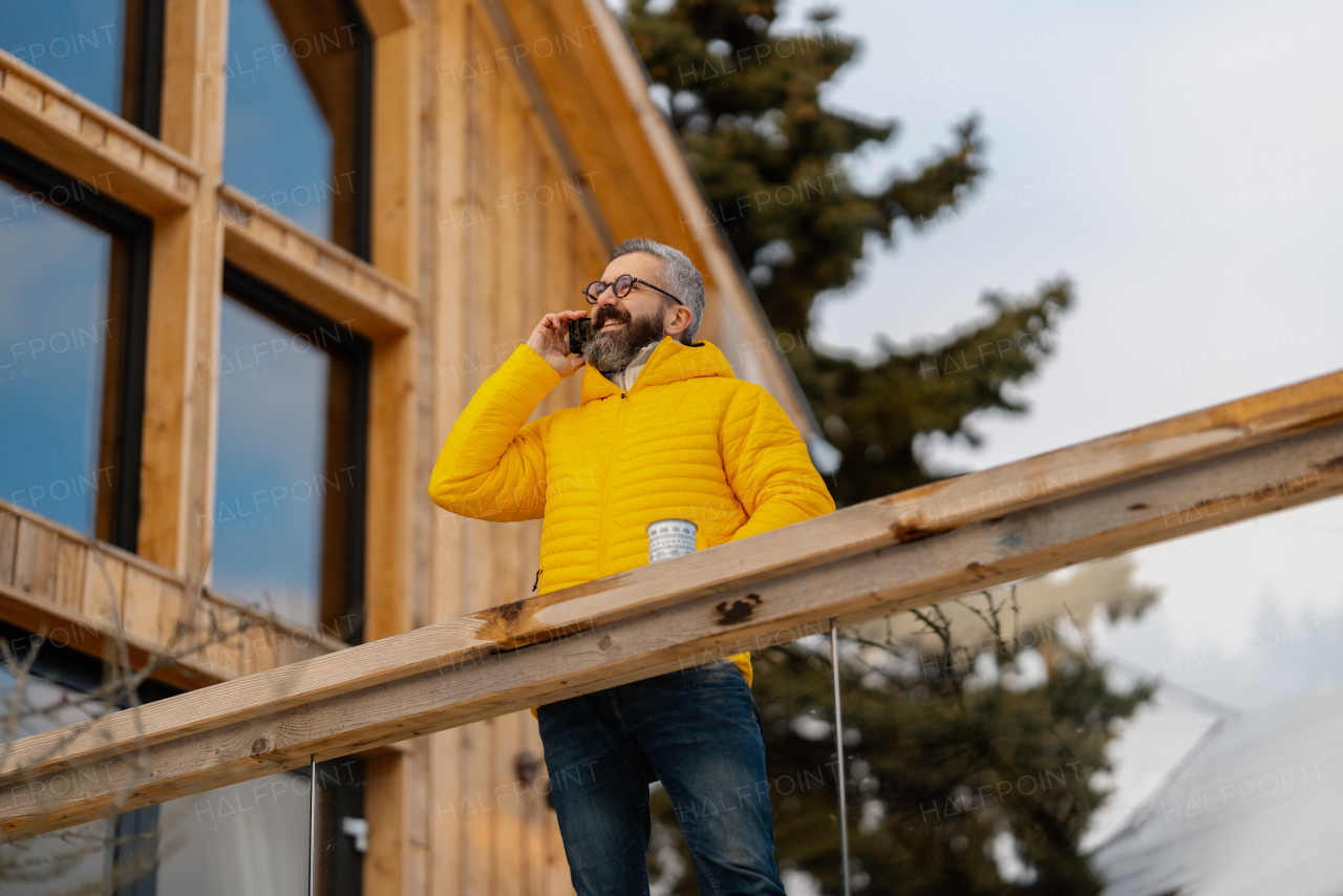 Mature man standing on cabins patio, drinking hot tea, coffee and enjoying beautiful winter day. Handsome man spending relaxing, stress-free winter weekend in cabin in mountains, enjoying alone time.