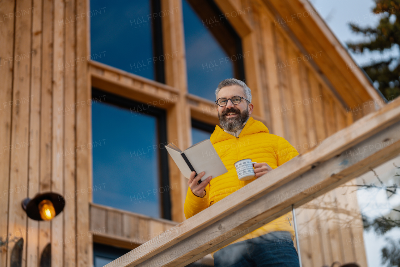 Mature man standing on cabins patio, drinking hot tea, coffee and reading book. Handsome man spending relaxing, stress-free winter weekend in cabin in mountains, enjoying alone time.