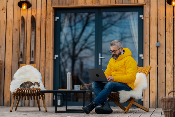 Man working from cozy cabin in mountains, sitting on terrace with laptop, enjoying cup of coffee, making call with client. Concept of remote work from beautiful, peaceful location. Hygge at work.