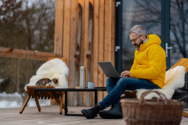 Man working from cozy cabin in mountains, sitting on terrace with laptop, enjoying cup of coffee. Concept of remote work from beautiful, peaceful location. Hygge at work.