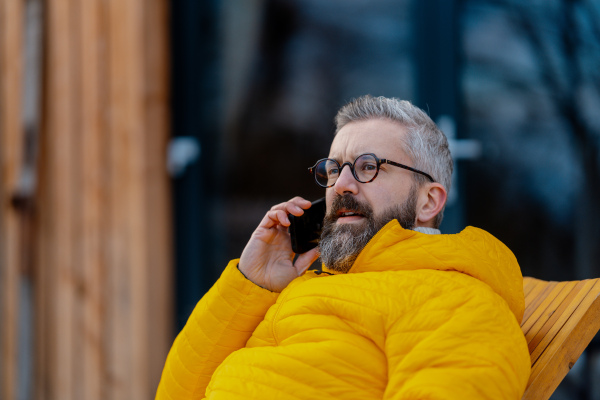 Portrait of handsome man phone calling from patio, during cold winter day. Handsome man spending relaxing, stress-free winter weekend in cabin in mountains, enjoying alone time.