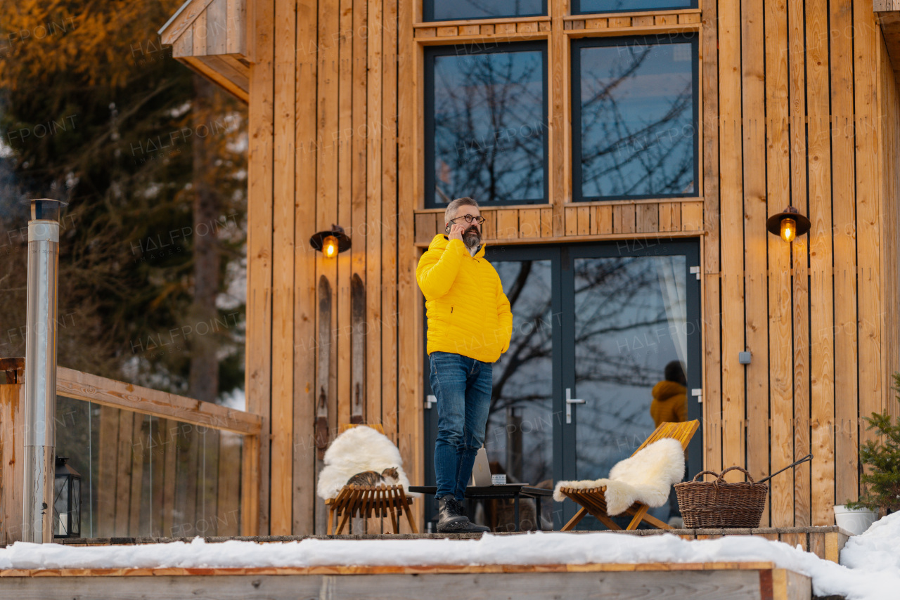 Mature man phone calling from patio, drinking hot tea, coffee and enjoying beautiful winter day. Handsome man spending relaxing, stress-free winter weekend in cabin in mountains, enjoying alone time.