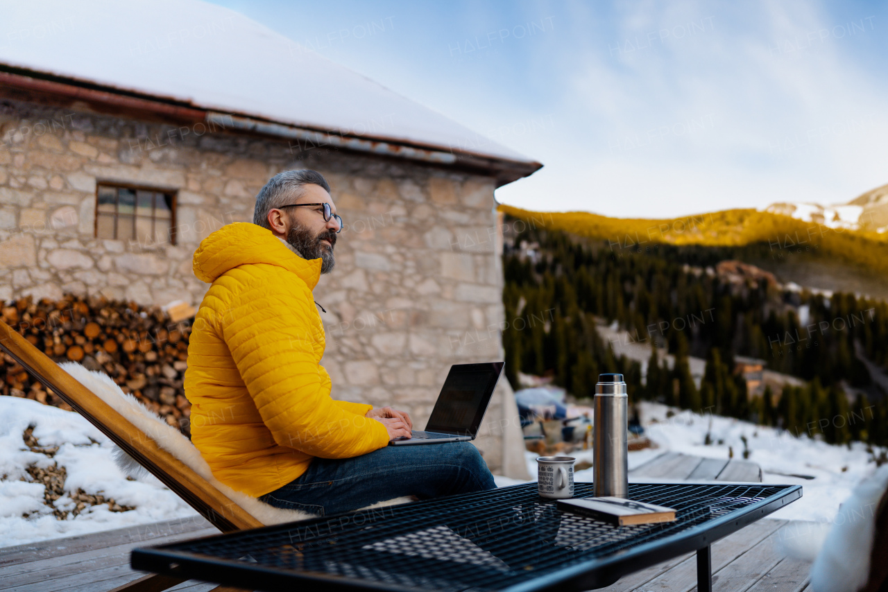 Man working from cozy cabin in mountains, sitting on terrace with laptop, enjoying cup of coffee. Concept of remote work from beautiful, peaceful location. Hygge at work.