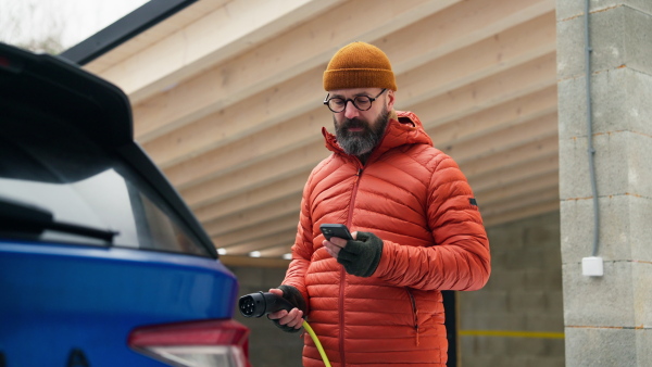 Mature man charging electric car during cold snowy day, using electric vehicle charging app, checking energy consumption, battery life on smart phone. Charging and driving electric vehicles during winter season.