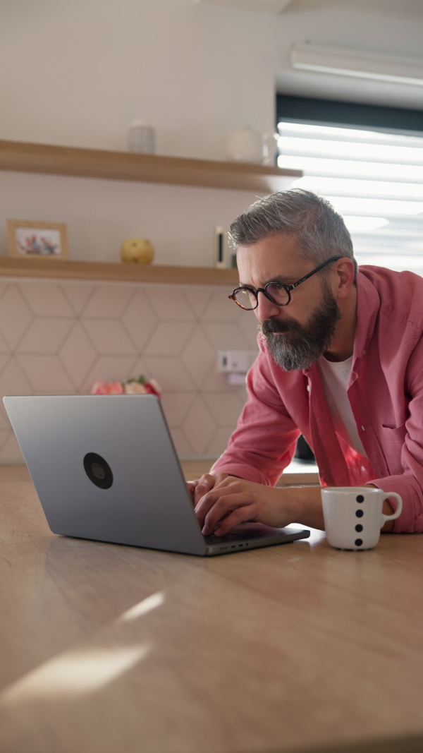 Mature man in pink shirt working from home on laptop, standing by kitchen island, with cup of coffee, holding smartphone. Concept of remote work from home. Hygge at work.