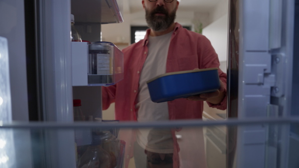 Handsome man putting lunchbox, takeaway container in fridge. Taking out lunch from fridge, eating leftovers. Do not trow food away, reducting food waste.