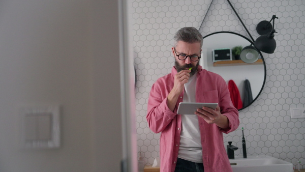 Man shopping online while brushing his teeth in bathroom. Holding tablet, scrolling and buying in eshop with toothbrush in mouth.