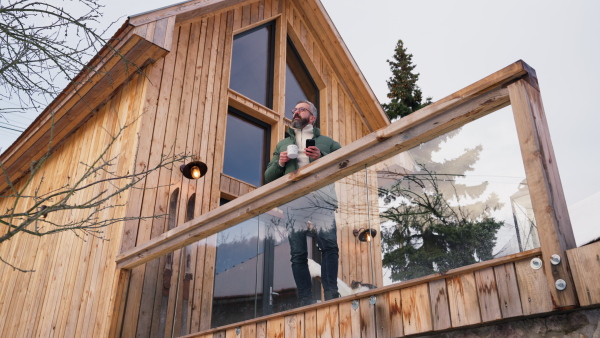 Mature man standing on cabins patio, drinking hot tea, coffee and enjoying beautiful winter day. Handsome man spending relaxing, stress-free winter weekend in cabin in mountains, enjoying alone time.