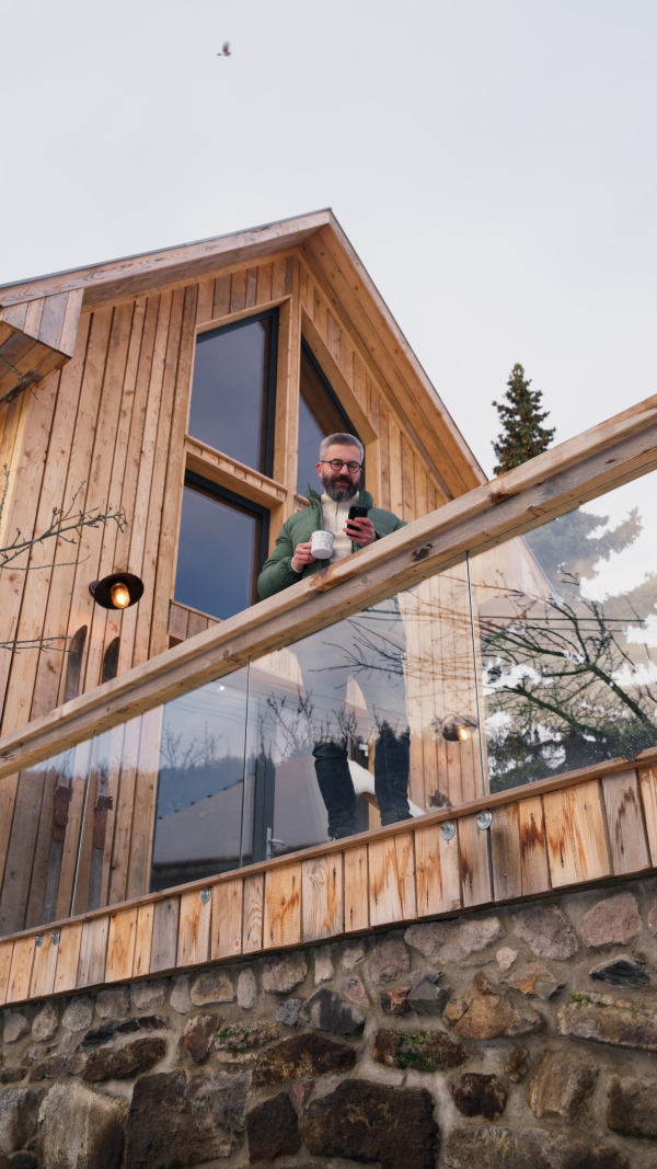 Mature man standing on cabins patio, drinking hot tea, coffee and enjoying beautiful winter day. Handsome man spending relaxing, stress-free winter weekend in cabin in mountains, enjoying alone time.