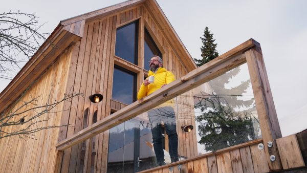 Mature man standing on cabins patio, drinking hot tea, coffee and enjoying beautiful winter day. Handsome man spending relaxing, stress-free winter weekend in cabin in mountains, enjoying alone time.