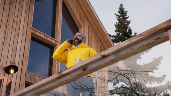 Mature man standing on cabins patio, drinking hot tea, coffee and enjoying beautiful winter day. Handsome man spending relaxing, stress-free winter weekend in cabin in mountains, enjoying alone time.