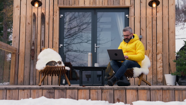 Man working from cozy cabin in mountains, sitting on terrace with laptop, enjoying cup of coffee. Concept of remote work from beautiful, peaceful location. Hygge at work.