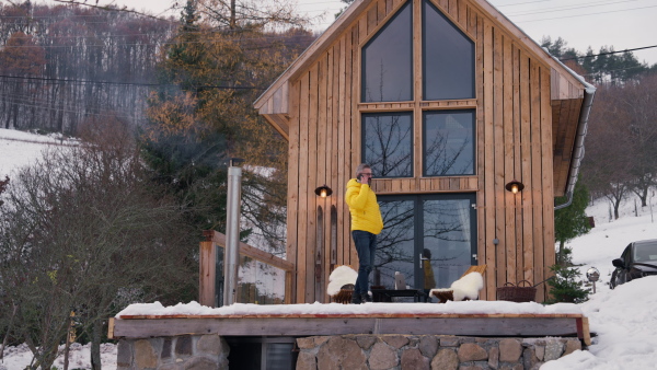 Mature man phone calling from patio, drinking hot tea, coffee and enjoying beautiful winter day. Handsome man spending relaxing, stress-free winter weekend in cabin in mountains, enjoying alone time.