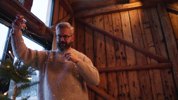 Mature man working setting christmas lights in wooden cabin. Concept of remote holidays.