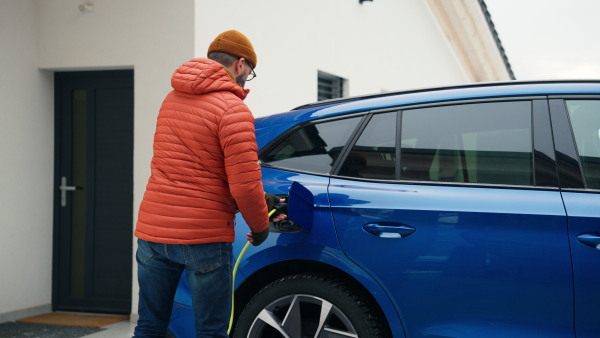 Man charging electric car during cold snowy day. Side view of hansome mature man putting charger in charging port during cold weather. Charging and driving electric vehicles during winter season.