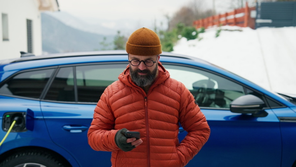 Mature man charging electric car during cold snowy day, using electric vehicle charging app, checking energy consumption, battery life on smart phone. Charging and driving electric vehicles during winter season.