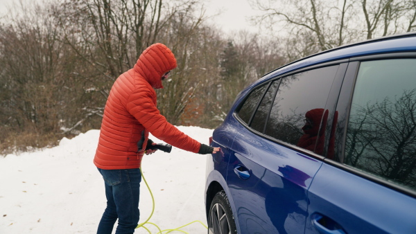 Man charging electric car during cold snowy day. Side view of hansome mature man putting charger in charging port during cold weather. Charging and driving electric vehicles during winter season.