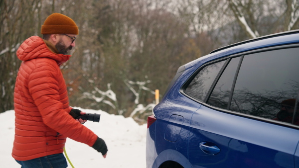 Man charging electric car during cold snowy day. Side view of hansome mature man putting charger in charging port during cold weather. Charging and driving electric vehicles during winter season.