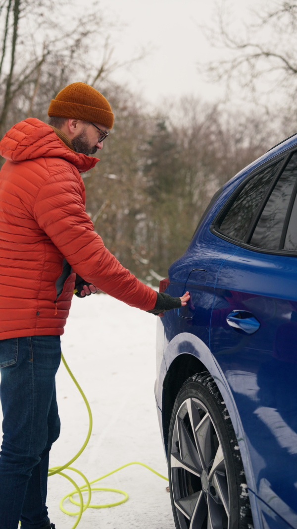 Man charging electric car during cold snowy day. Side view of hansome mature man putting charger in charging port during cold weather. Charging and driving electric vehicles during winter season.