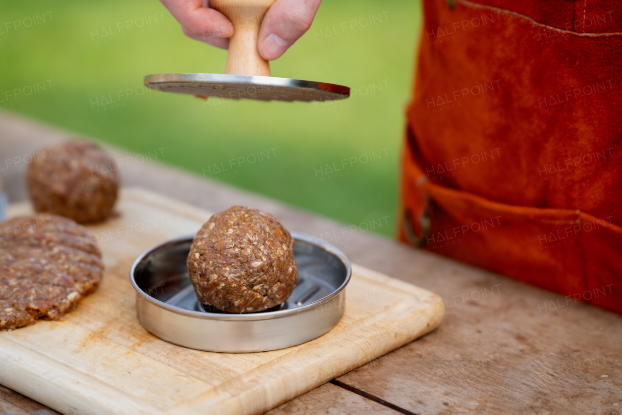 Man in apron preparing meat for hamburgers, standing outdoors. Using burger press to shape groud meat.