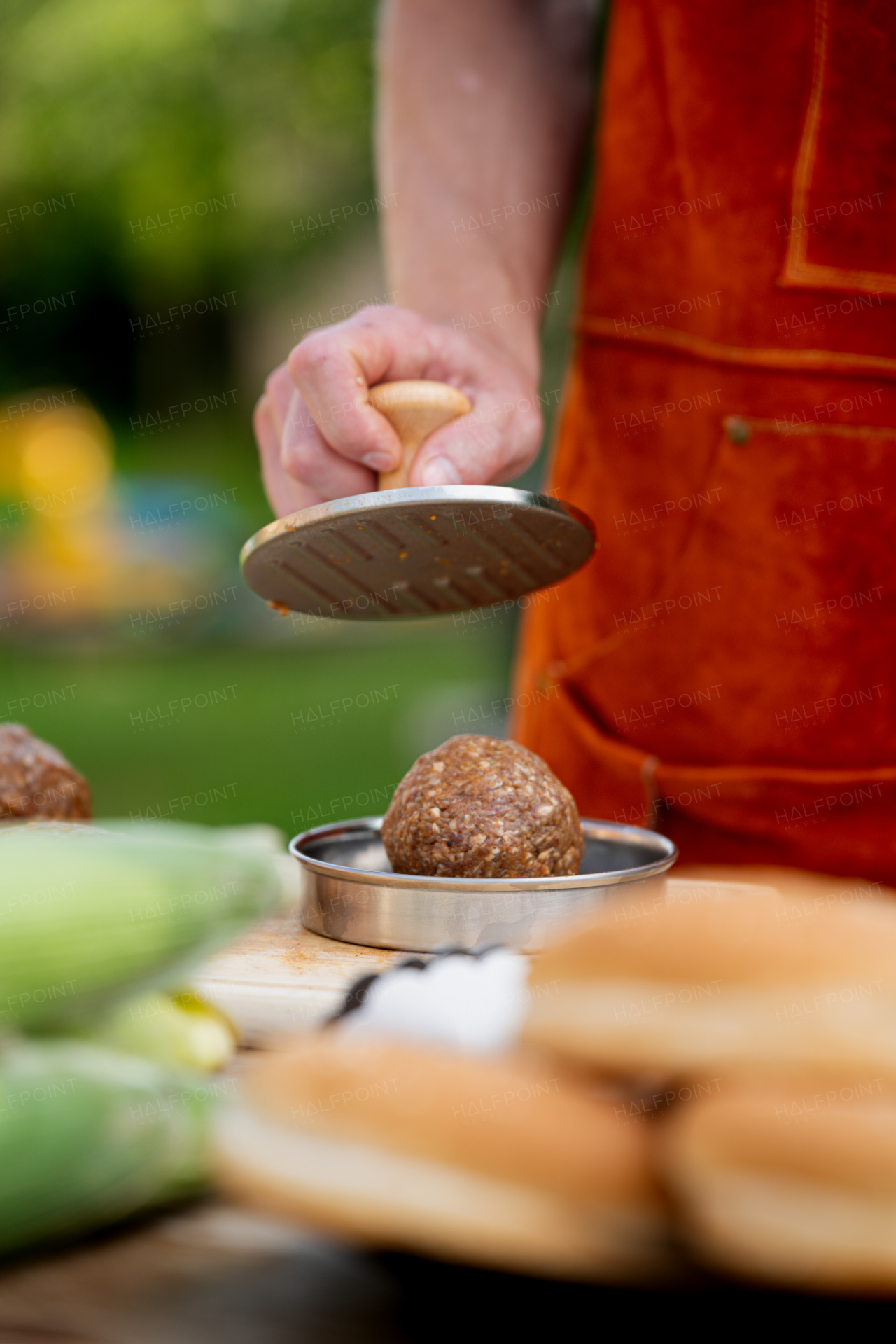 Man in apron preparing meat for hamburgers, standing outdoors. Using burger press to shape groud meat.