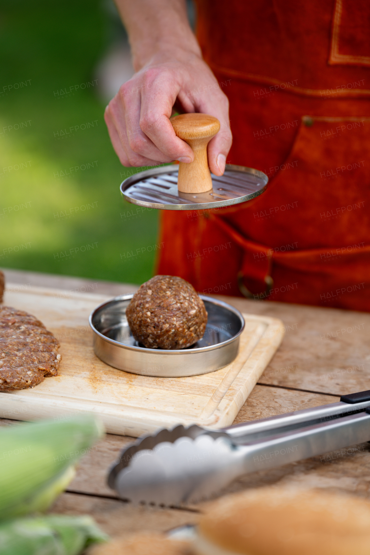 Man in apron preparing meat for hamburgers, standing outdoors. Using burger press to shape groud meat.