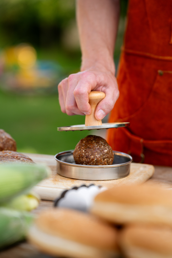 Man in apron preparing meat for hamburgers, standing outdoors. Using burger press to shape groud meat.