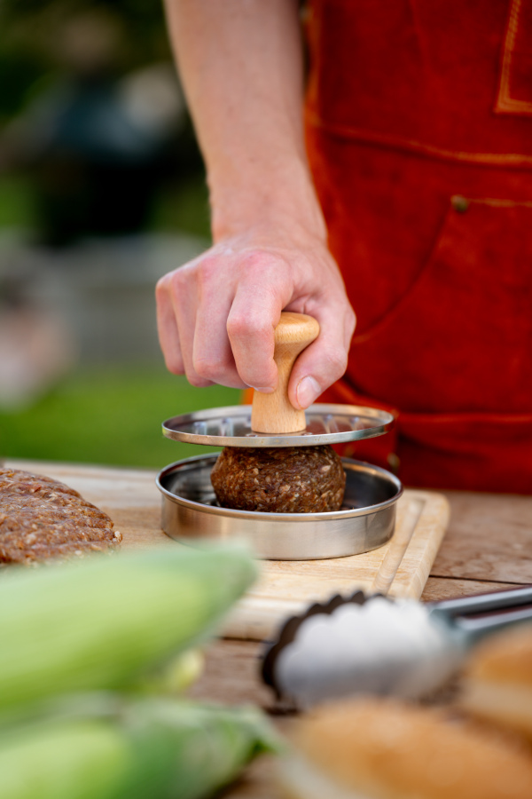 Man in apron preparing meat for hamburgers, standing outdoors. Using burger press to shape groud meat.