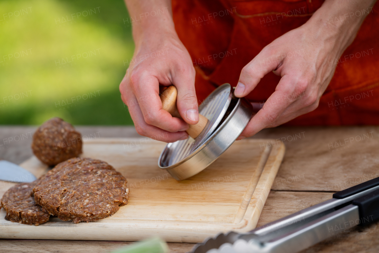 Man in apron preparing meat for hamburgers, standing outdoors. Using burger press to shape groud meat.