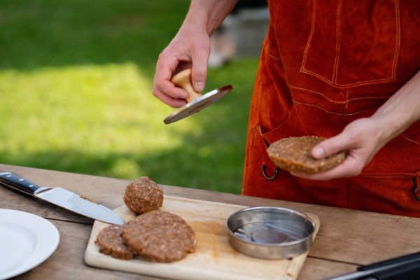 Man in apron preparing meat for hamburgers, standing outdoors. Using burger press to shape groud meat.